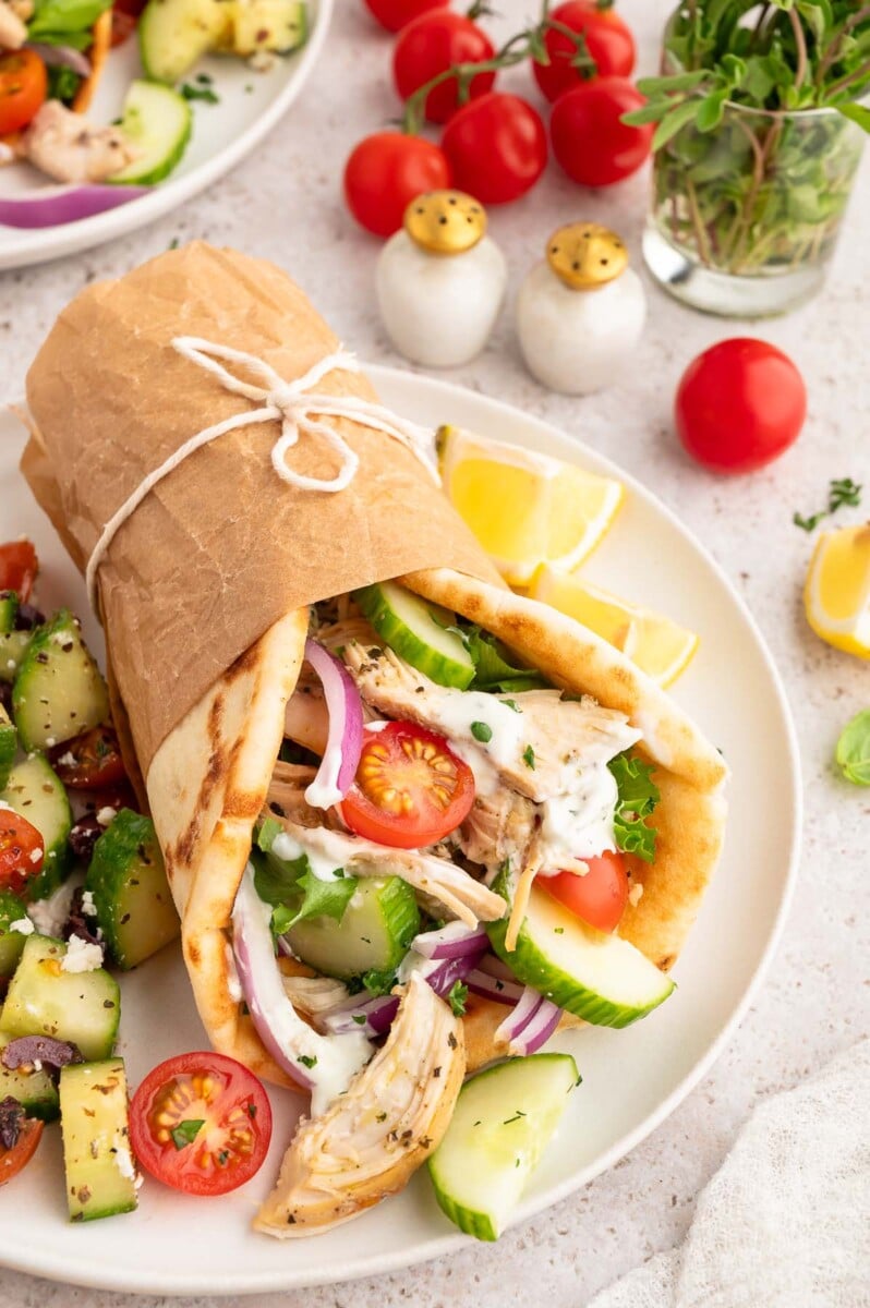 Greek Shredded chicken on a pita with cherry tomatoes and cucumbers wrapped in paper with a greek salad on the side.