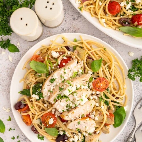 A plate of Greek spaghetti topped with chicken and a plate of spaghetti on the side.