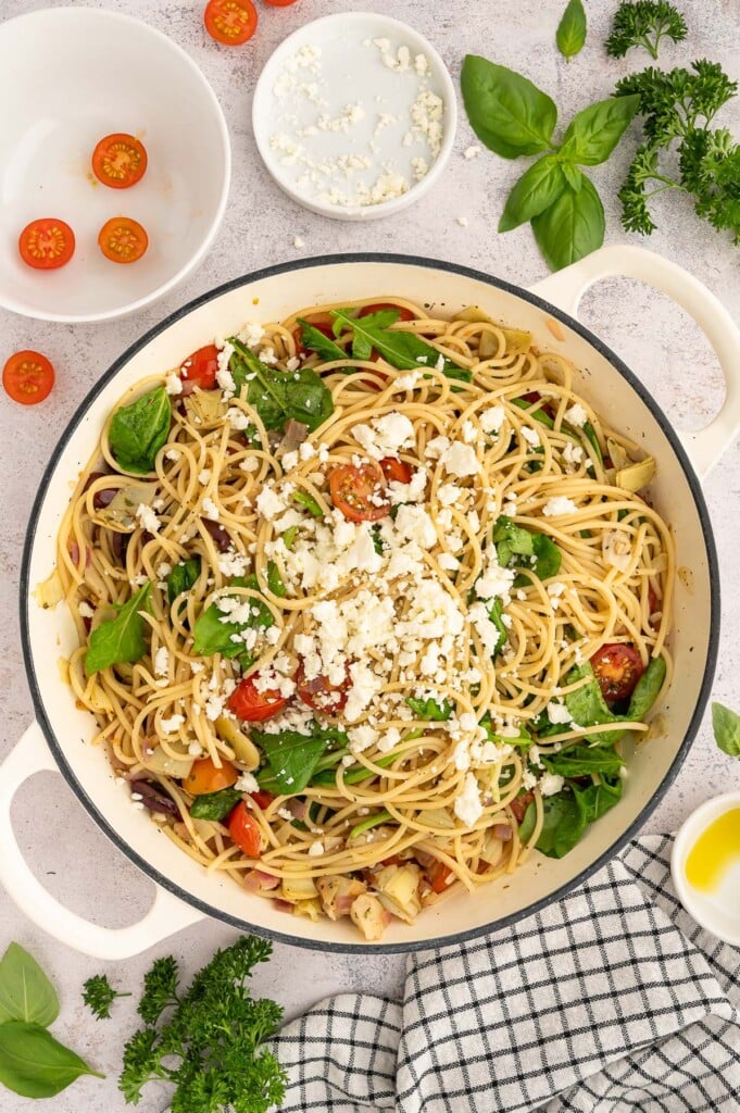 A dutch oven full of greek spaghetti with more ingredients on the side.