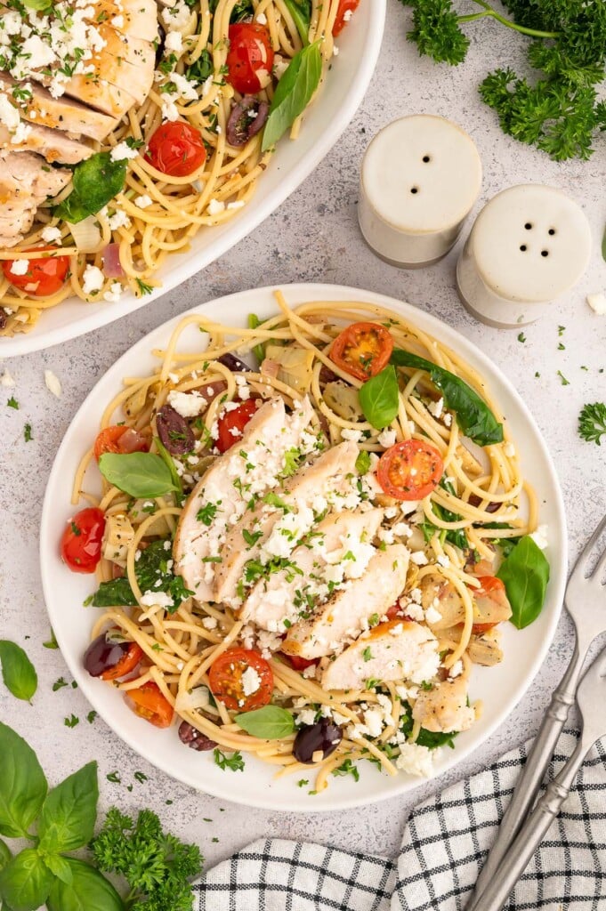 Greek spaghetti topped with sliced chicken on a white plate and a plate of more greek pasta on the side.