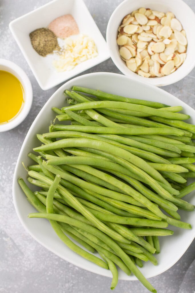 Ingredients for green beans almondine laid out on a grey background in small white bowls