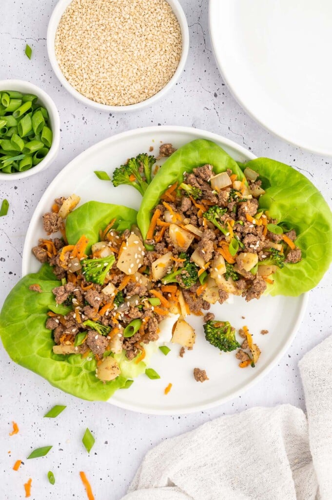 Two Ground Beef Lettuce Wraps on a white plate with bowls of green onions and dipping sauce on the side