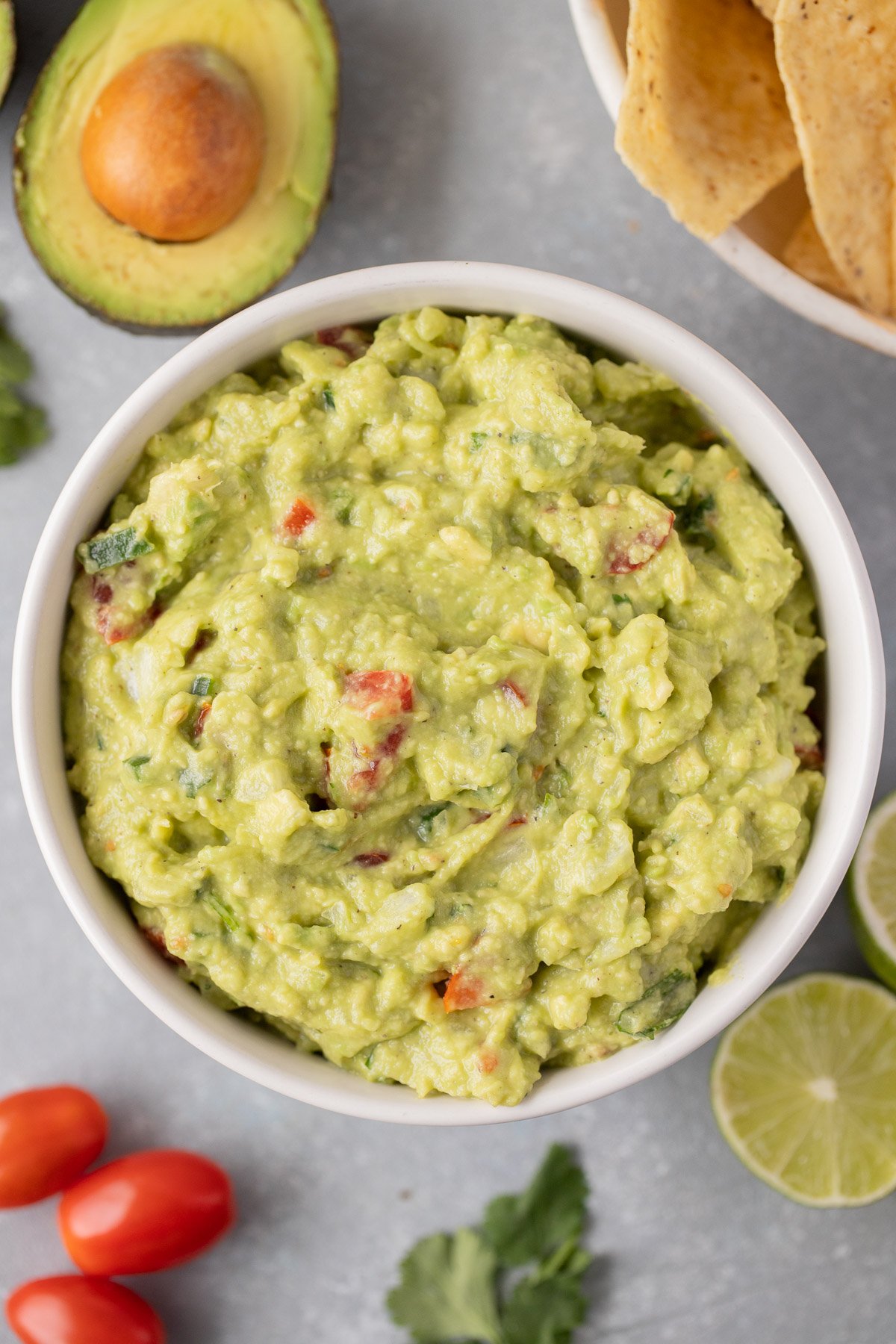 An overhead shot of the best guacamole in a white bowl with avocados, tomatoes, lime, and cilantro on the side