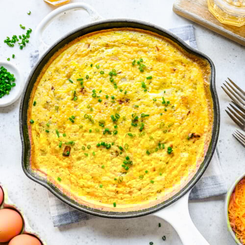 An overhead shot of a cast iron pan of ham and cheese frittata topped with fresh herbs on a white background
