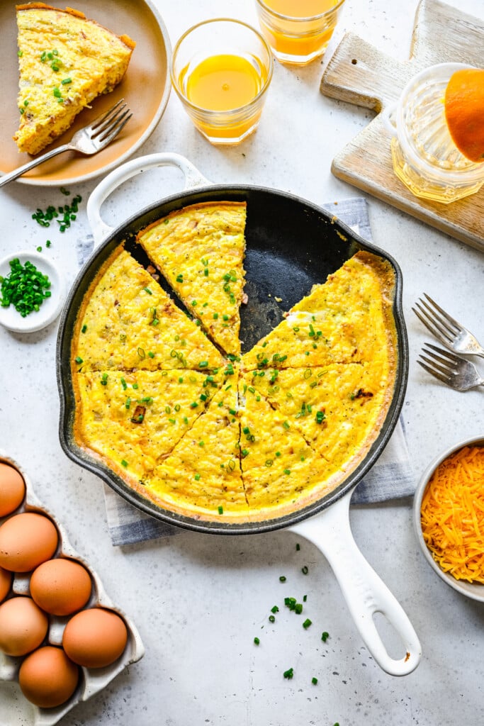 An overhead shot of a cast iron pan of ham and cheese frittata topped with fresh herbs with half cut and one piece taken out on a white background