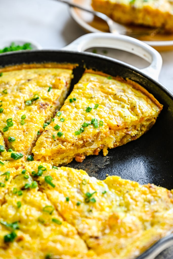 A cast iron pan of ham and cheese frittata with a piece taken out topped with fresh chives
