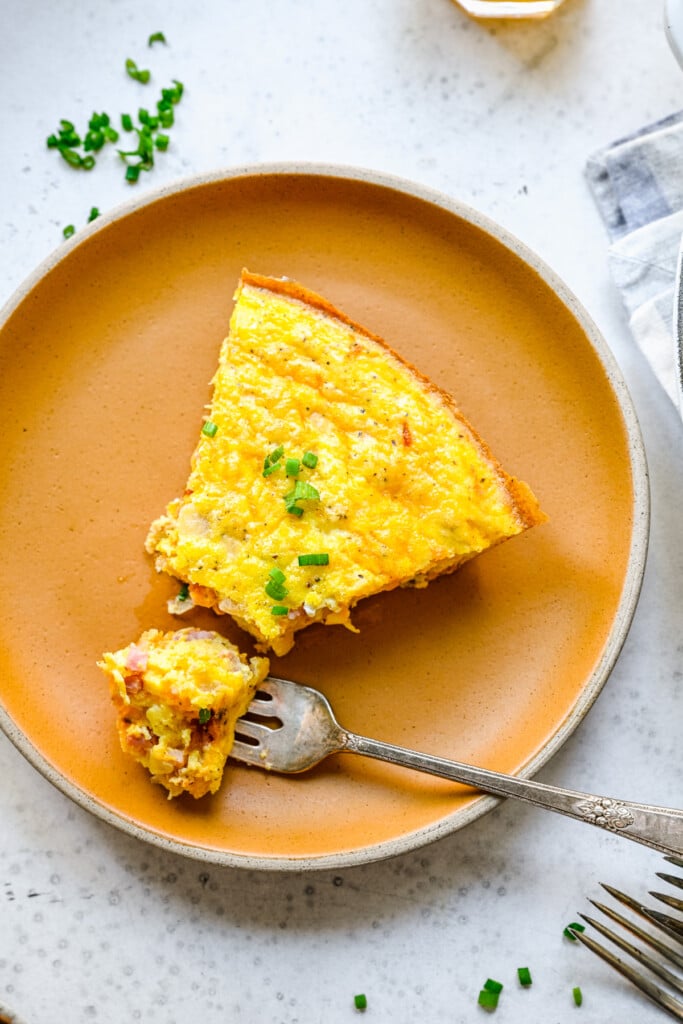 A piece of ham and cheese frittata with a bite taken out with a fork on a brown plate on a white background