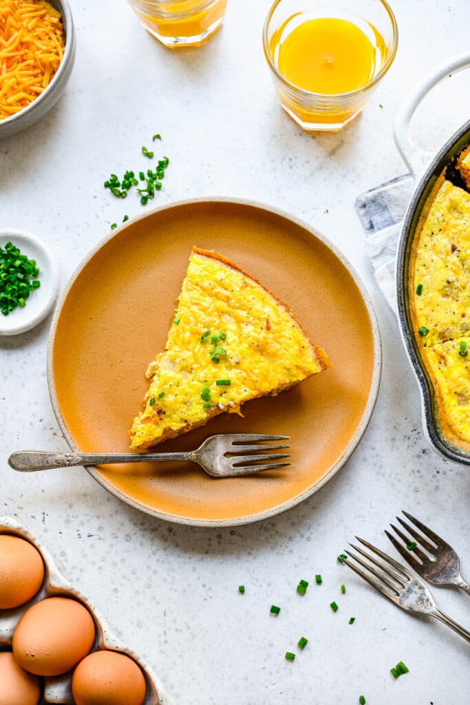 A piece of keto frittata on a brown plate on a white background with a cast iron pan and eggs on the side