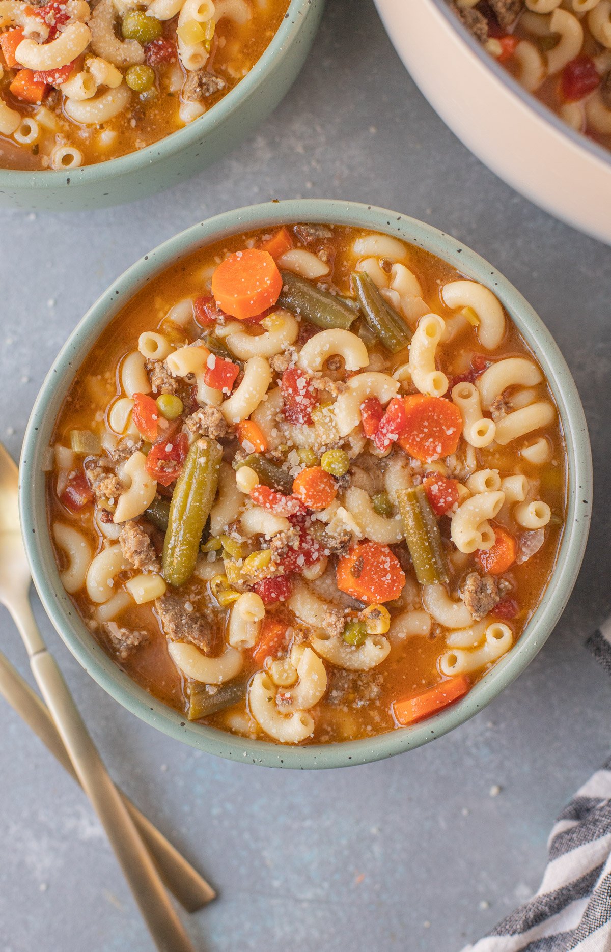 An overhead shot of a bowl of hamburger vegetable soup on a grey background with gold spoons on the side