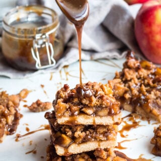 A stack of Healthy Apple Crumble Bars with caramel being poured on top of it. Apples in the background