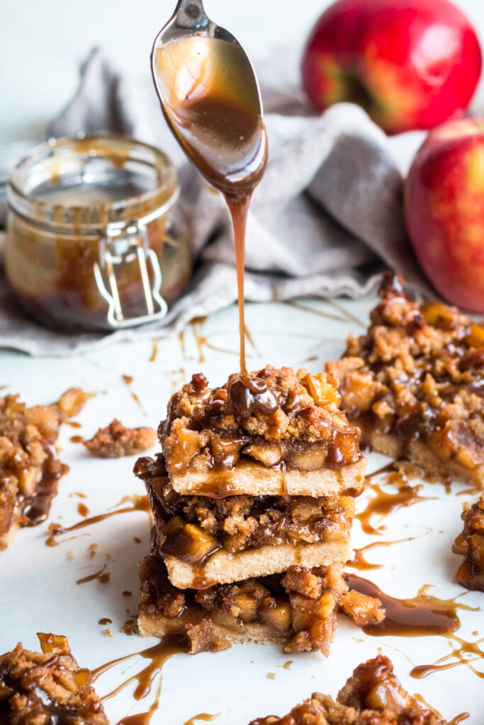 A stack of Healthy Apple Crumble Bars with caramel being poured on top of it. Apples in the background