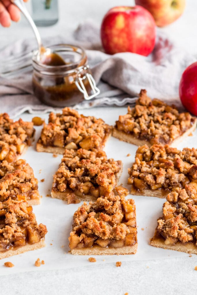 Gluten Free Apple Crumble Bars on a white background with fruit and caramel off to the side.