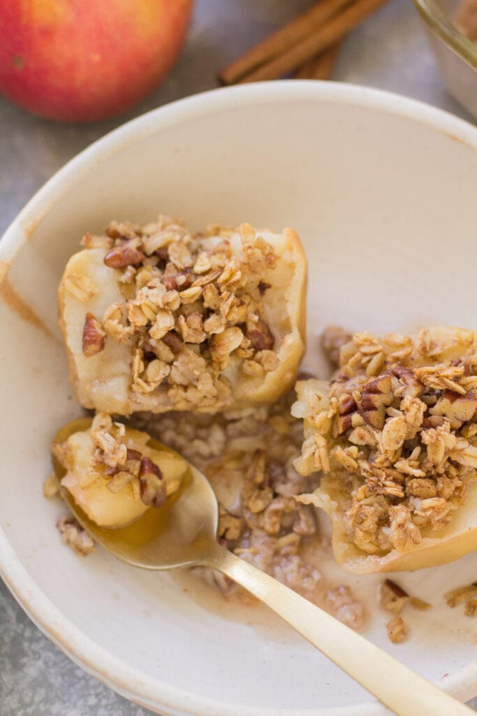Healthy Baked Apples cut in half in a shallow white bowl. A gold spoon is off to the side scooping a bite out of the apple