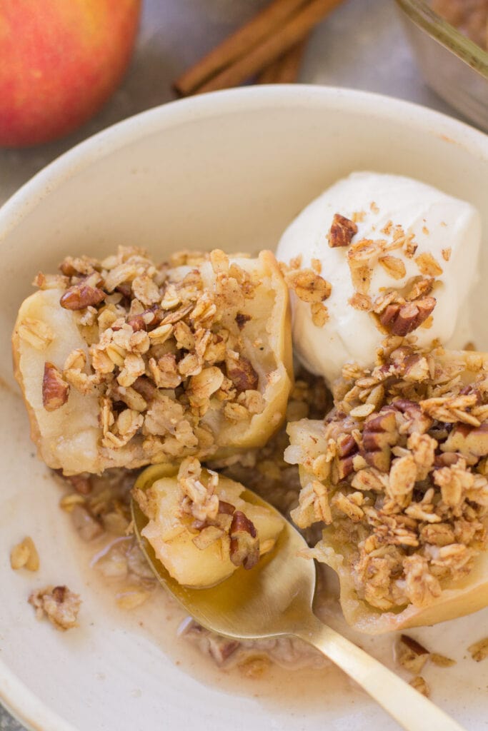 A baked apple cut in half in a white bowl. Ice cream is off to the side in the bowl + there are apples+ a blue linen off to the side.