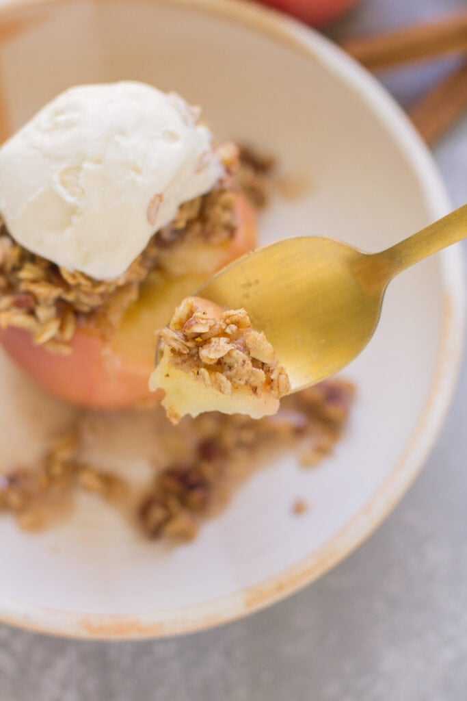 A gold spoon taking a bite out of a healthy baked apple in a white bowl on a blue background