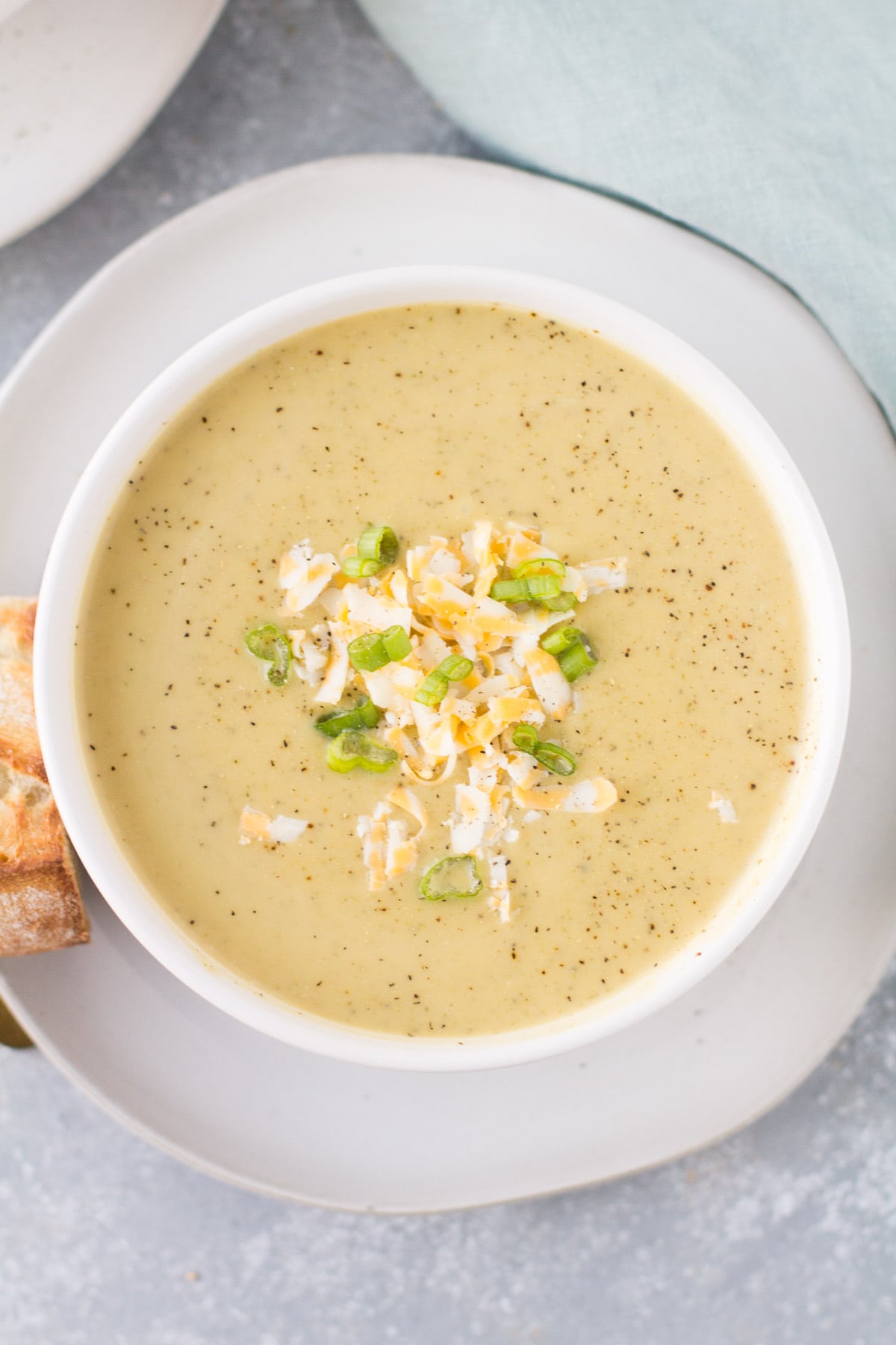 An overhead shot of Healthy Broccoli Soup in a white bowl on a white plate with bread off to the side.