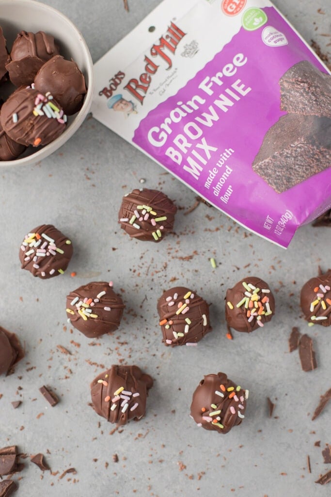 Brownie truffles on a grey background with a bowl of truffles, a bag of brownie mix + chocolate scattered around it.