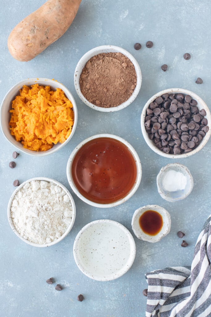 Ingredients for healthy sweet potato brownies in white bowls: mashed sweet potato, coconut oil, eggs, maple syrup, oat flour, vanilla extract, cocoa powder, baking soda, baking powder, chocolate chips 