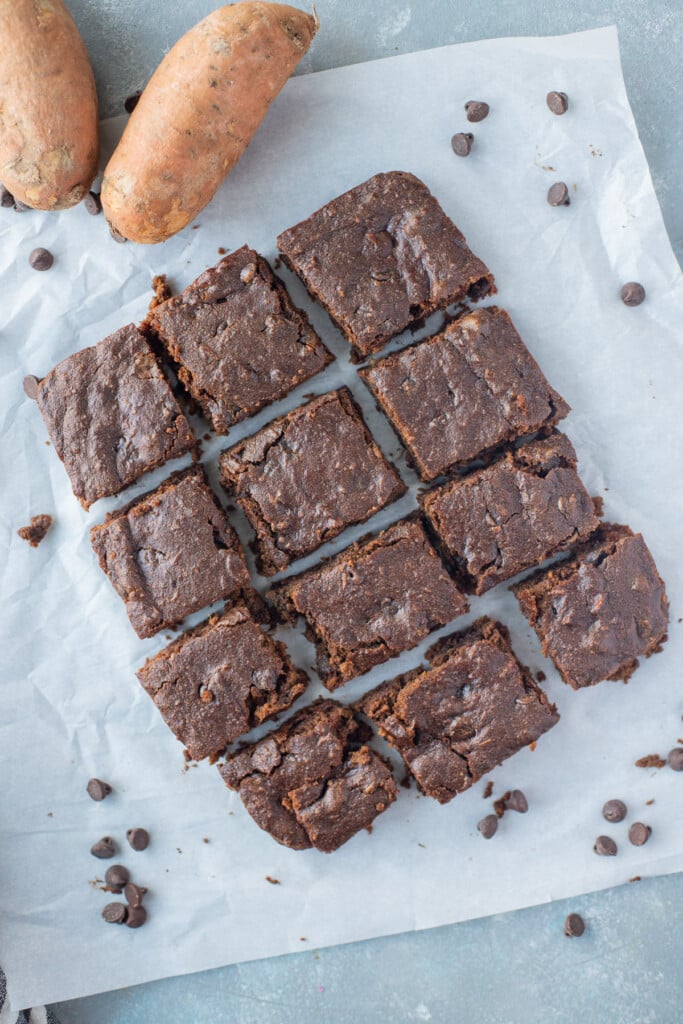 12 sweet potato brownie squares on a piece of parchment paper with chocolate chips and sweet potatoes on the side on a grey background