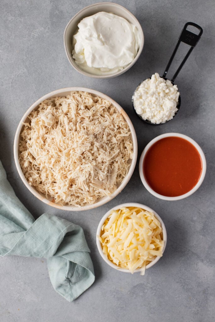 Ingredients for healthy buffalo chicken dip laid out on a blue background with a striped linen