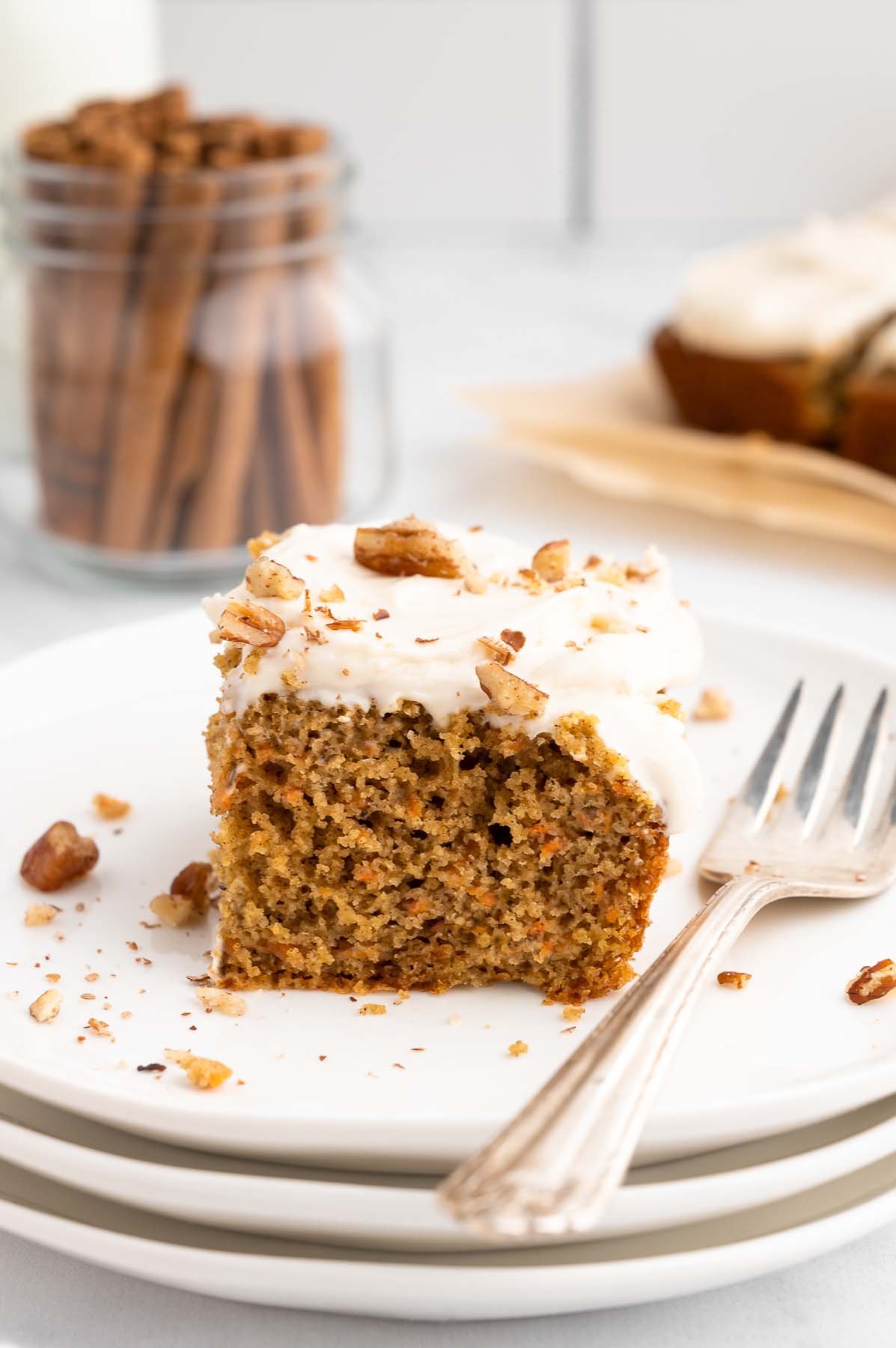 A piece of healthy carrot cake with healthy cream cheese frosting on a white plate with a fork on the side