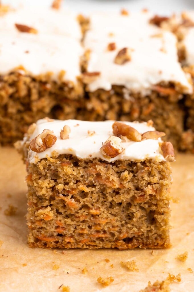 A piece of healthy carrot cake topped with healthy cream cheese frosting with the rest of the cake in the background on a piece of parchment paper