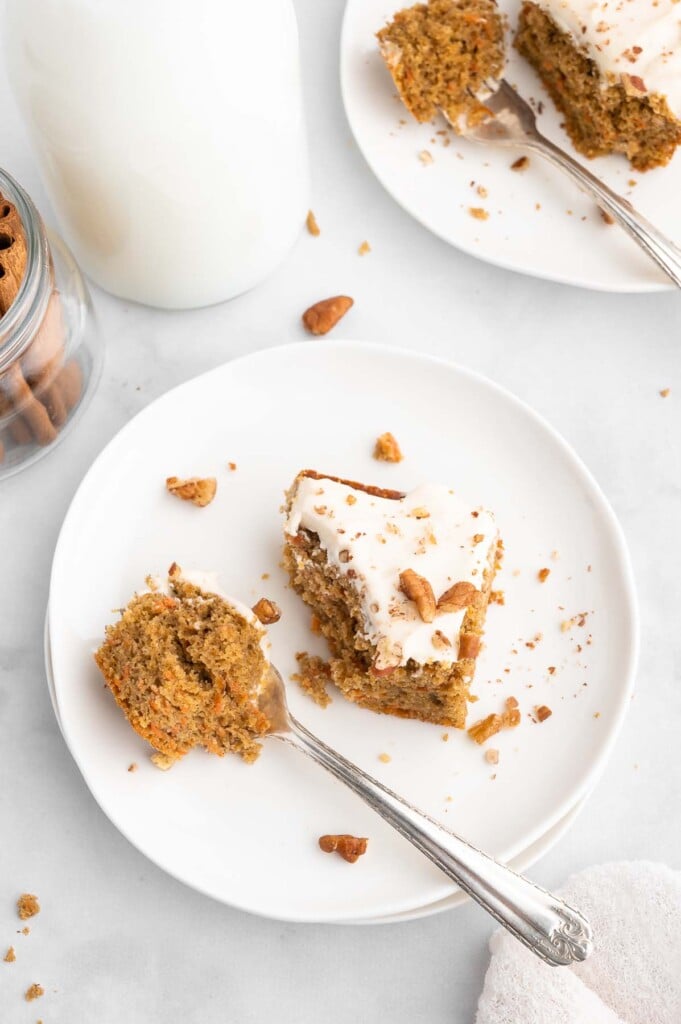 An overhead shot of a piece of healthy carrot cake on a white plate with a silver fork taking a bite out of it on a white background