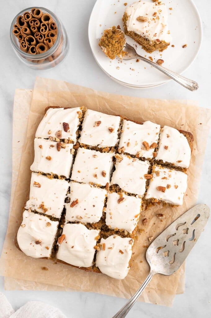 An overhead shot of healthy carrot cake sliced into squares with two pieces missing on a piece of parchment paper and a plate on the side