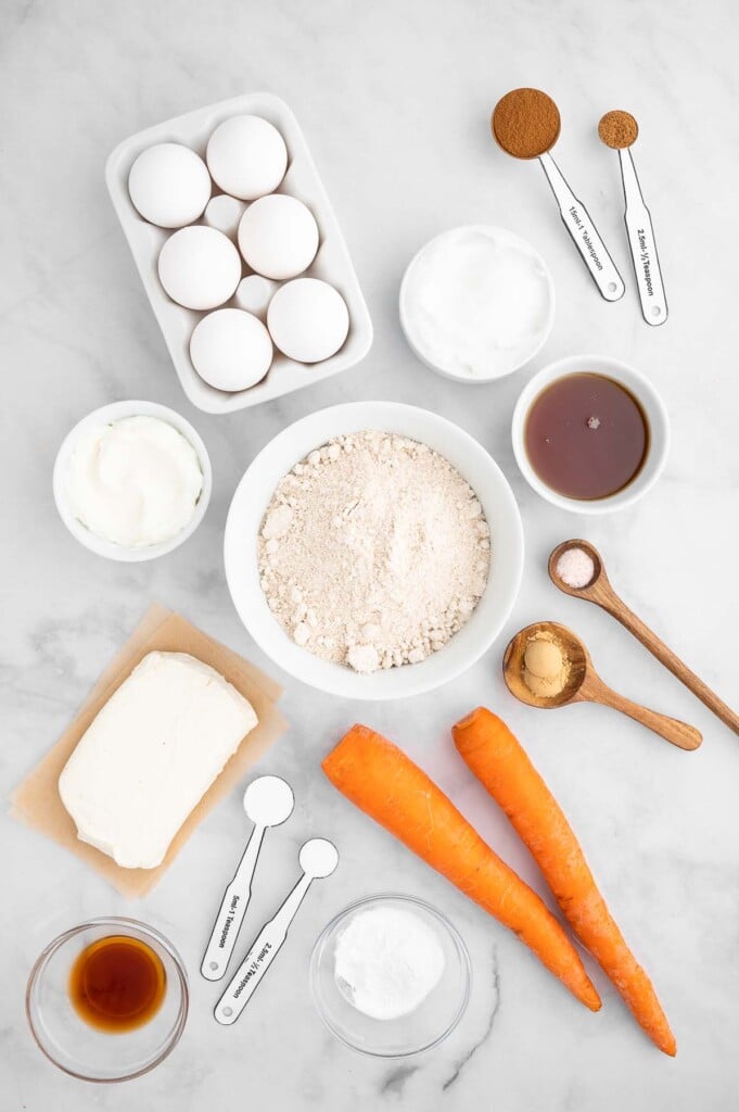 Ingredients for healthy carrot cake in white bowls: large carrots, eggs, maple syrup, coconut oil, greek yogurt, vanilla extract, oat flour, baking powder, baking soda, ground cinnamon, ground ginger, ground nutmeg, sea salt, neufchatel cheese, greek yogurt, maple syrup, sea salt