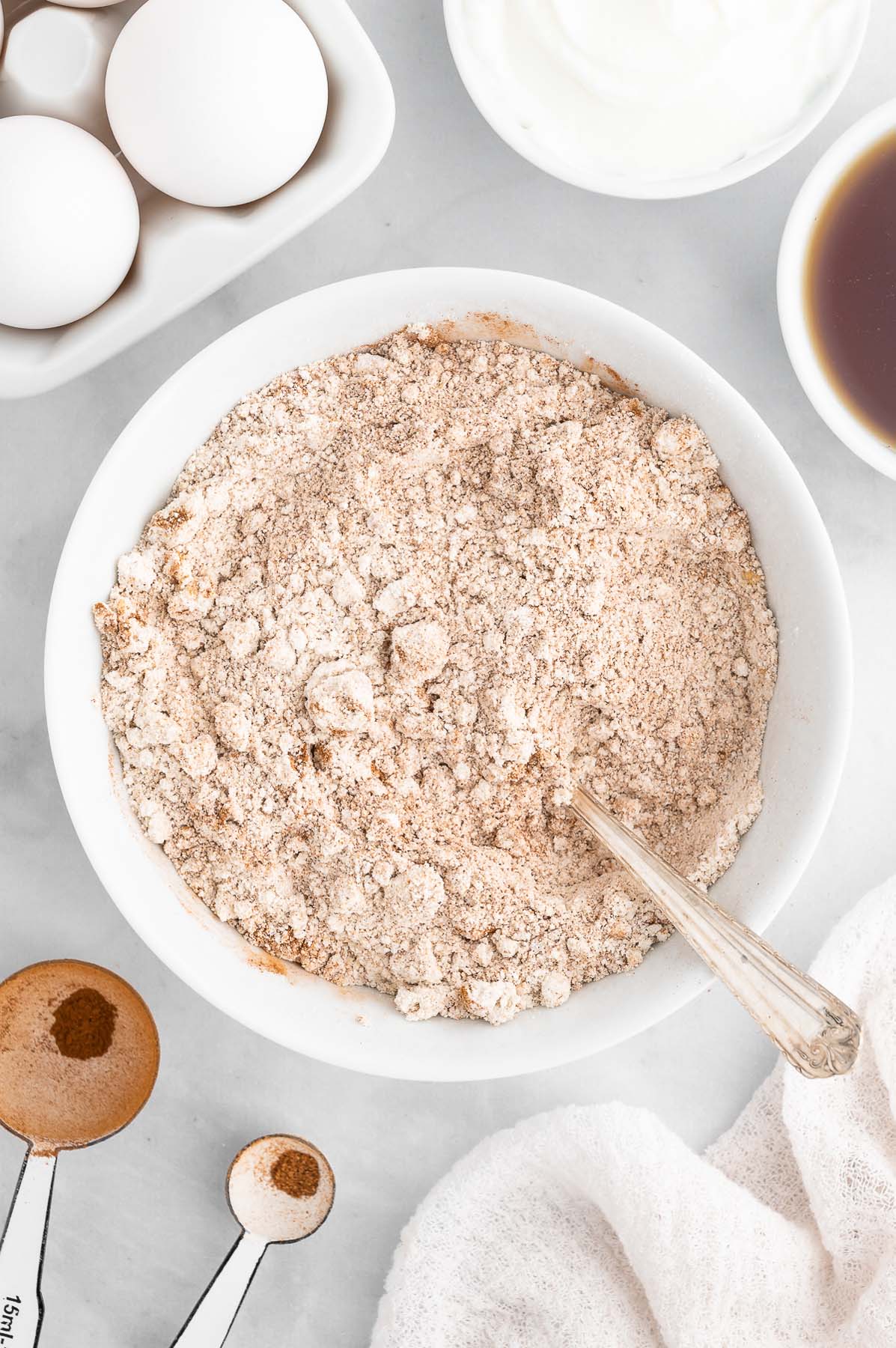 Dry ingredients for healthy carrot cake in a white bowl after mixing together with bowls, eggs, and measuring spoons on the side