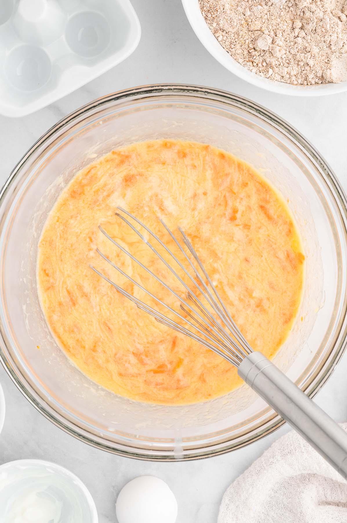Wet ingredients for healthy carrot cake being whisked together in a clear bowl on a marble background