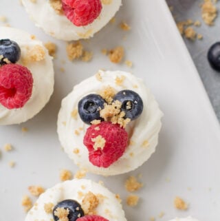 An overhead shot of mini healthy cheesecakes on a white square plate with berries and crumbs off to the side
