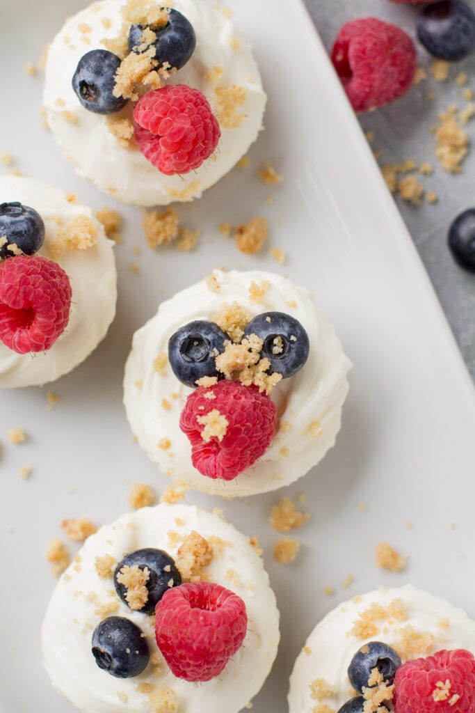 An overhead shot of mini healthy cheesecakes on a white square plate with berries and crumbs off to the side