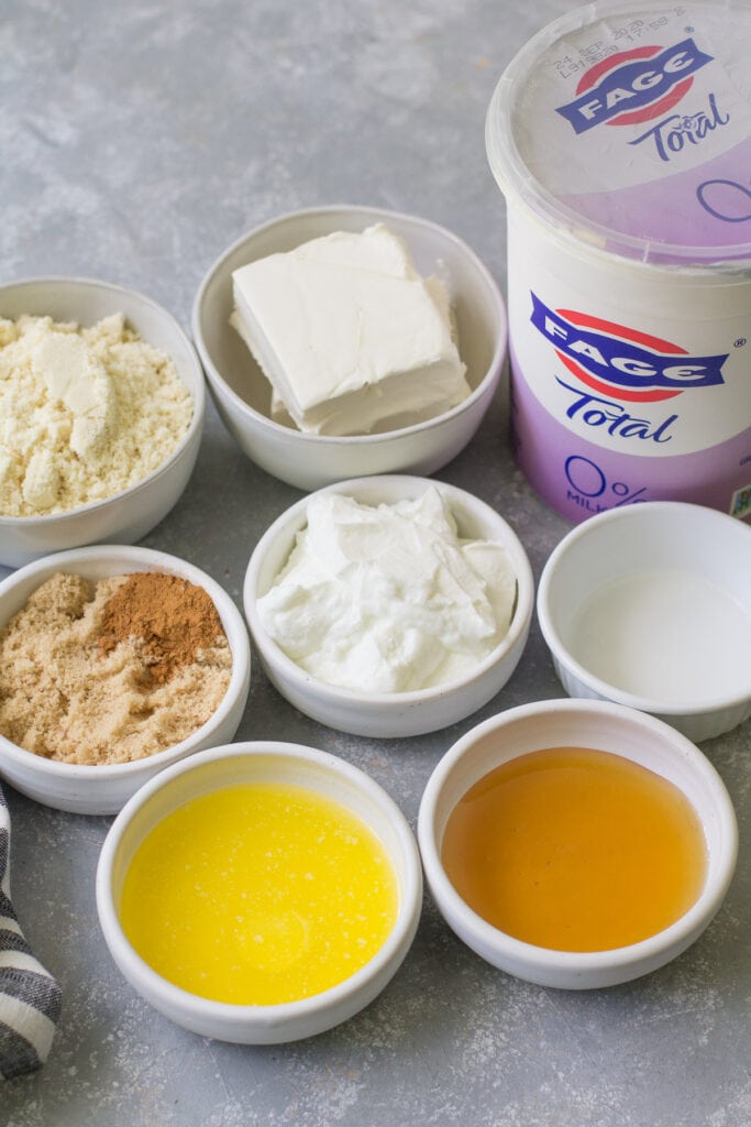 Ingredients for healthy cheesecakes in small white bowls: almond flour, cream cheese, yogurt, vanilla extract, honey, butter, brown sugar and cinnamon