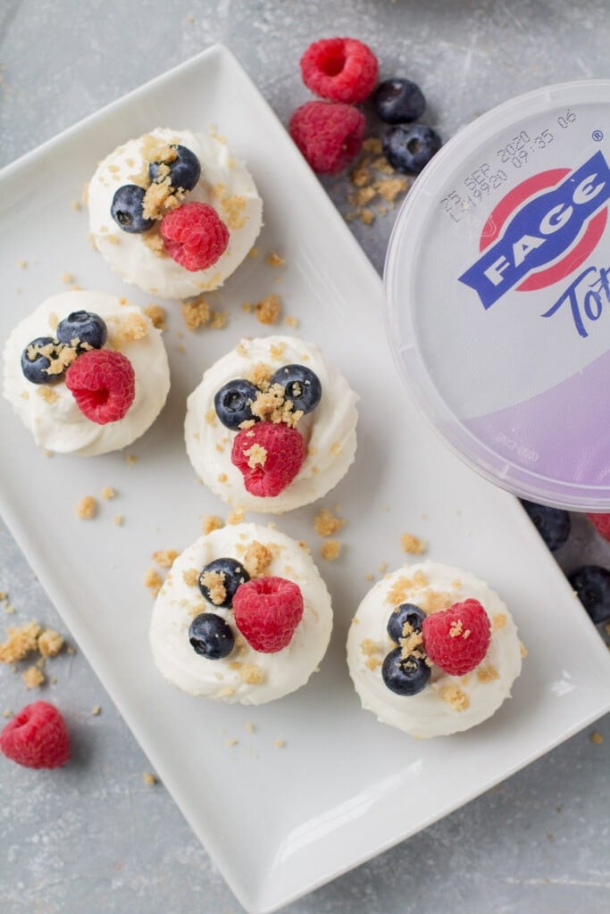 An overhead shot of mini greek yogurt cheesecakes on a rectangle white plate with a container of yogurt off to the side.