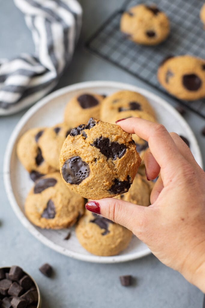 A hand holding up a single healthy chocolate chip cookie with a plate of cookies and cookies on a wire baking rack in the background