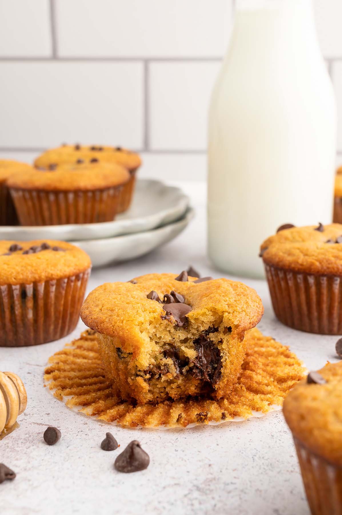 A Healthy Chocolate Chip Muffin with a bite taken out of it sitting on it's cupcake liner with more cupcakes scattered around on a white counter.
