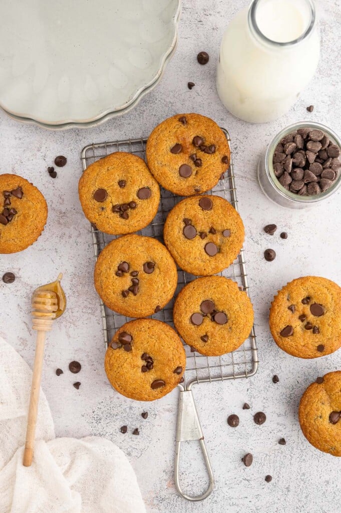 6 Healthy Chocolate Chip Muffins on a wire rack with chocolate chips and more muffins scattered around with a glass jar of milk on the side. 