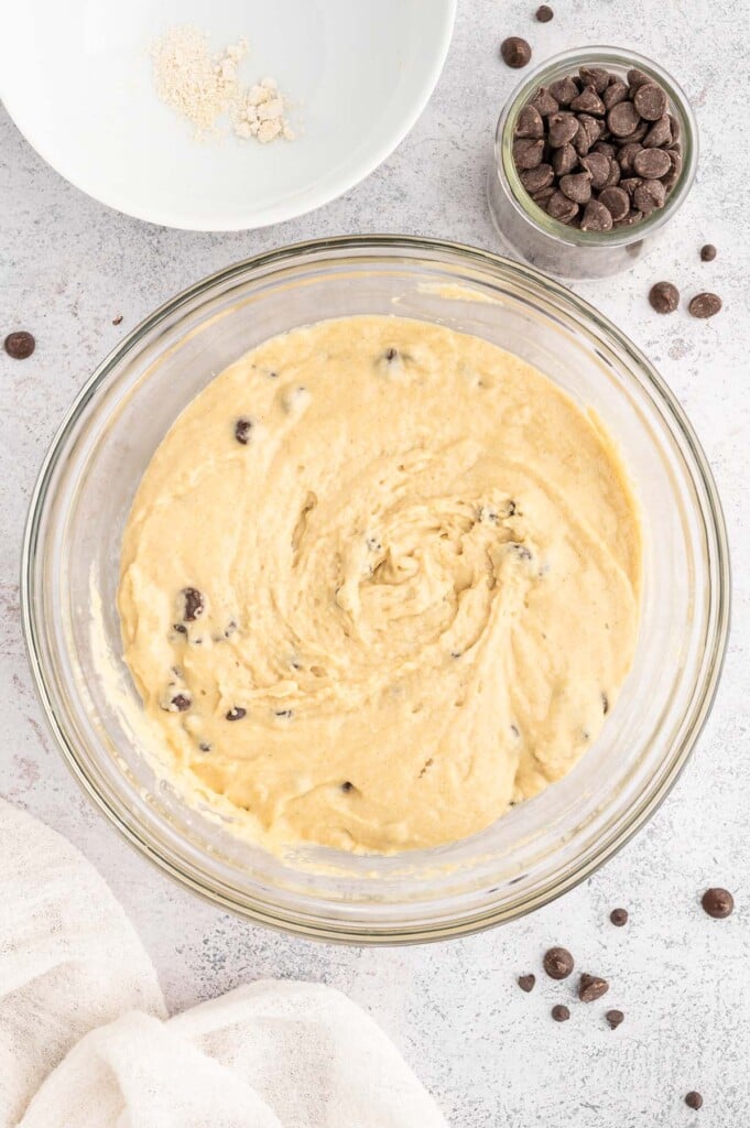 Chocolate Chip Muffin Batter in a clear glass bowl with more chocolate chips on the side on a white background.