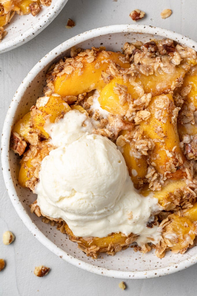 A close up peach crisp in a white speckled bowl with a scoop of ice cream on top