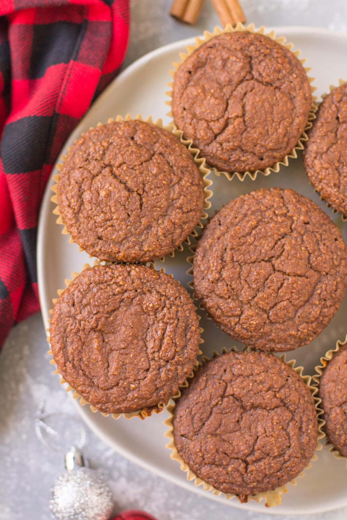 A white plate with healthy gingerbread muffins stacked on it. A red checked linen, cinnamon sticks + ornaments are scattered in the background