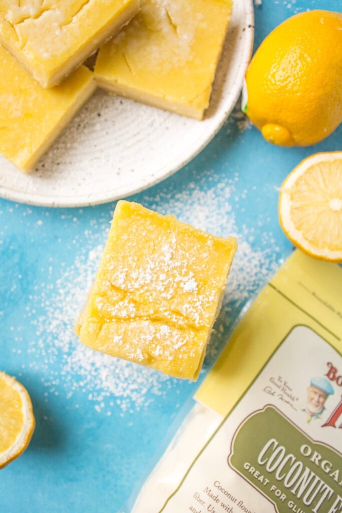 Healthy Lemon Bars stacked on a blue background with lemon bars on a white plate off to the side and a bag of coconut flour.