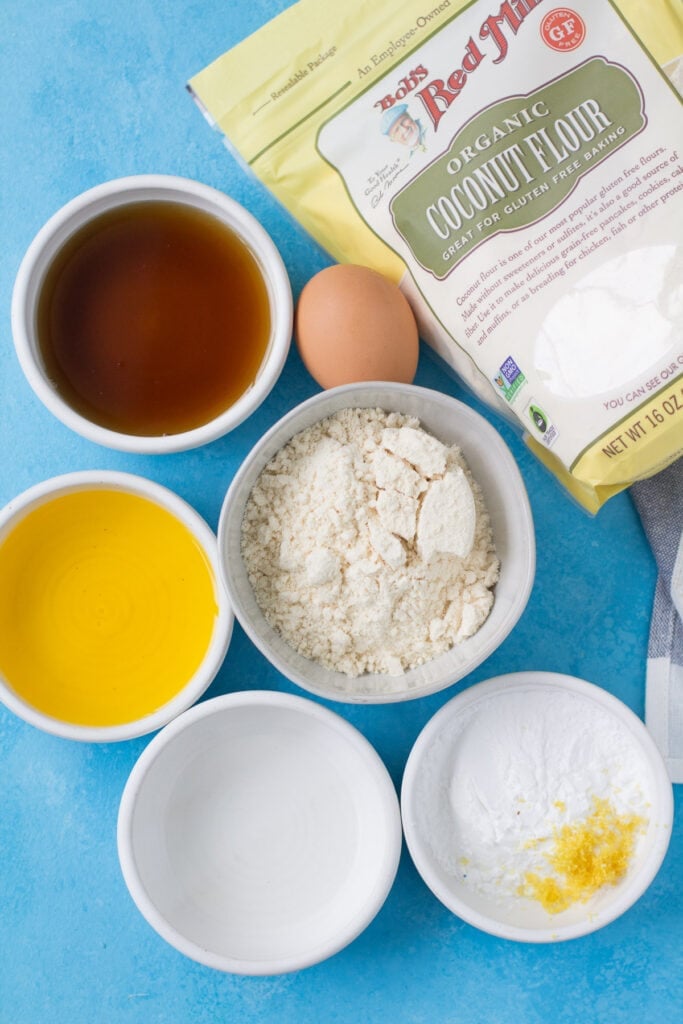 Ingredients for healthy lemon bars in small white bowls on a bright blue background - maple syrup, lemons, egg, melted ghee, coconut flour, tapioca flour.
