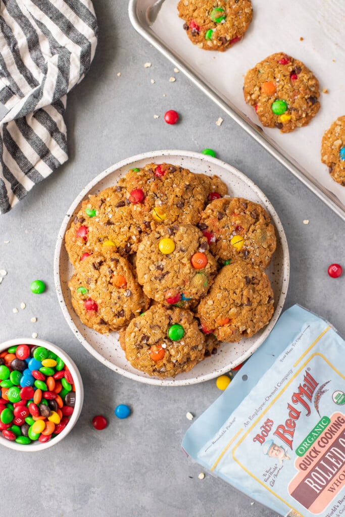 An overhead shot of a white plate filled with healthy monster cookies and m&ms, a baking sheet of cookies, and bob's red mill rolled oats on the side