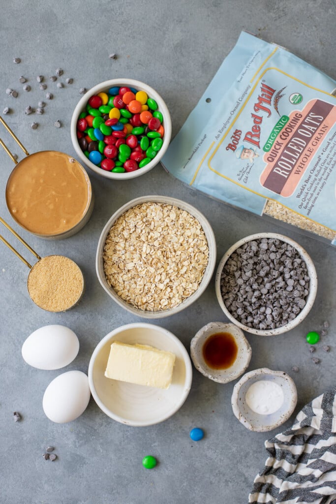 Ingredients for healthy monster cookies in white bowls: salted butter, brown sugar, creamy peanut butter, eggs, vanilla extract, baking soda, Bob's Red Mill Organic Quick Cook Oats, mini chocolate chips, m&ms