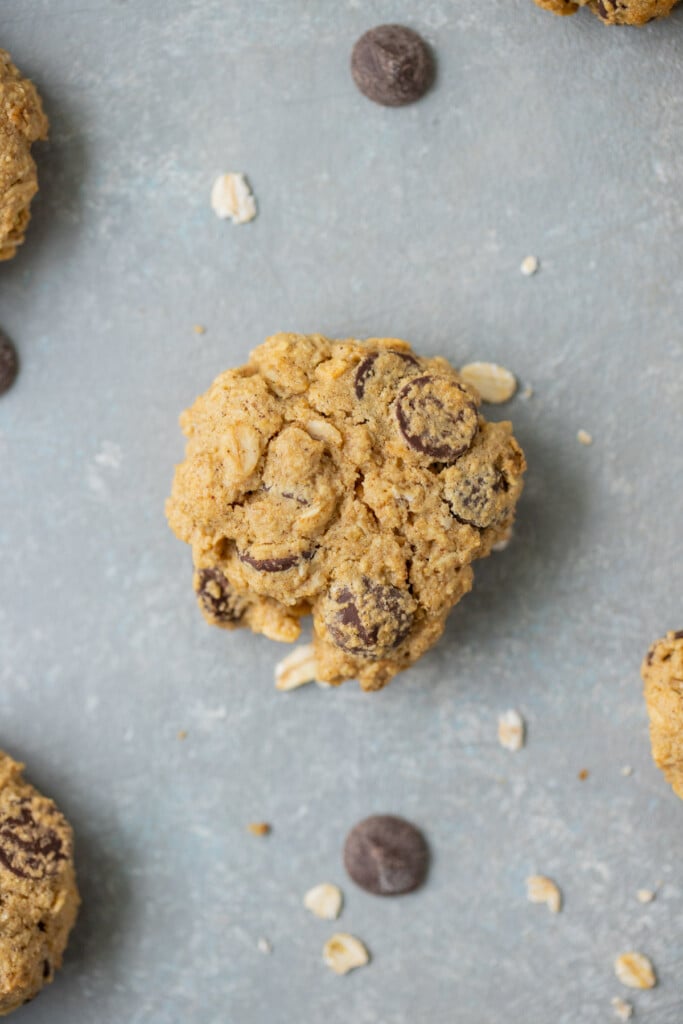 A healthy oatmeal chocolate chip cookie on a grey counter with more cookies, chocolate chips, and oats on the side
