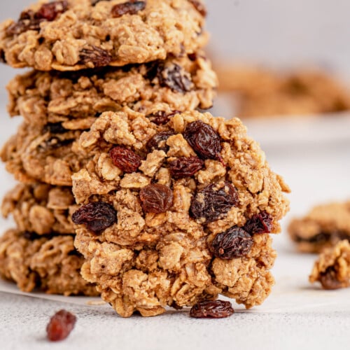 A oatmeal raisin cookie leaning on a stack of cookies on a grey background