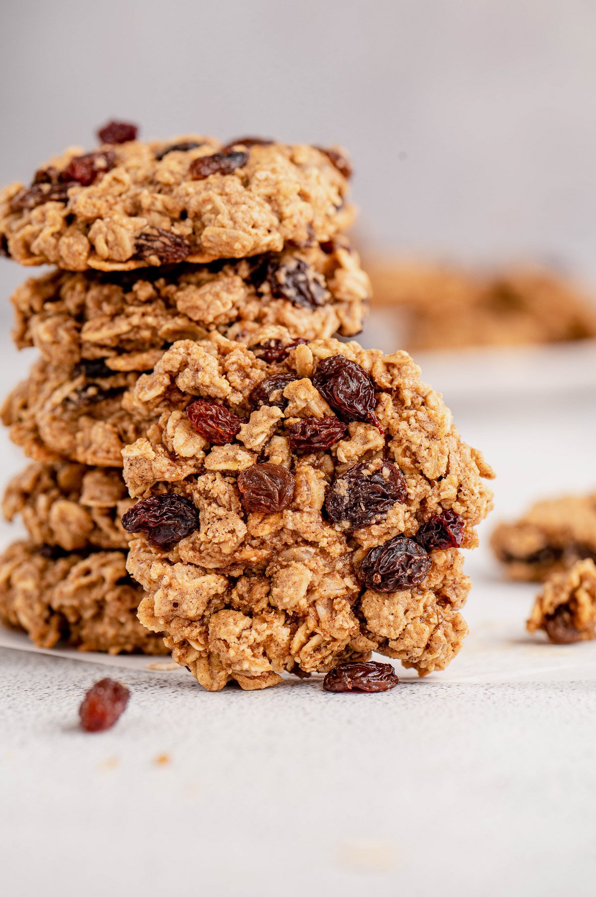 A oatmeal raisin cookie leaning on a stack of cookies on a grey background