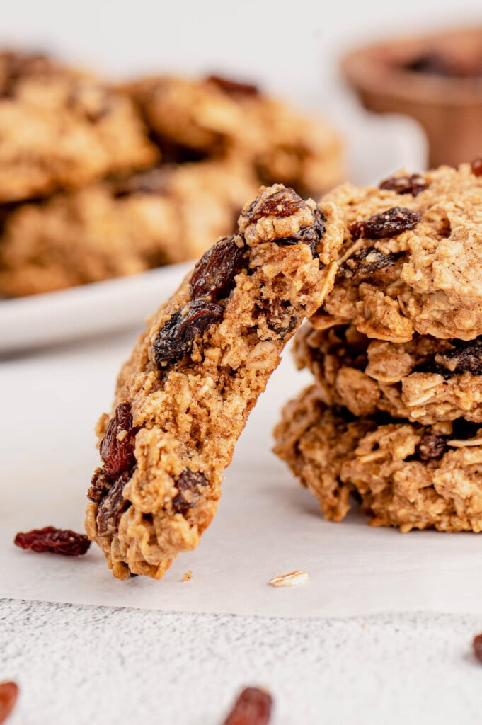An oatmeal raisin cookie leaning up against a stack of cookies with a plate of healthy oatmeal raisin cookies in the background