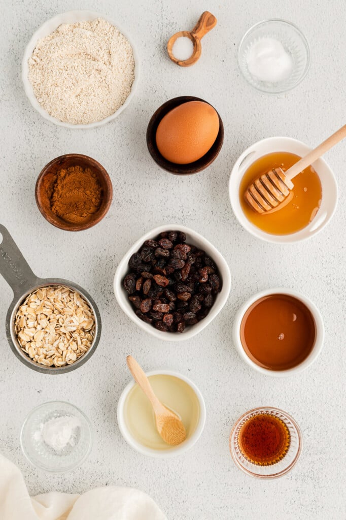 Ingredients for oatmeal raisin cookies in white bowls: raisins, rolled oats, oat flour, baking soda, powder, salt, cinnamon, coconut oil, maple syrup, honey, eggs, vanilla extract 